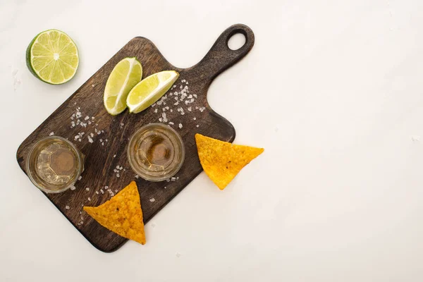 Top view of golden tequila with lime, salt and nachos near wooden cutting board on white marble surface — Stock Photo