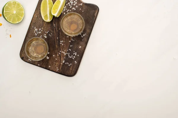 Blick von oben auf goldenen Tequila mit Kalk, Salz auf Holzschneidebrett auf weißem Marmor — Stockfoto