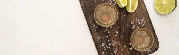 Вид сверху на золотую текилу с лаймом, соль на деревянной доске для резки на поверхности белого мрамора, панорамный снимок — стоковое фото