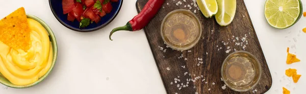 Top view of golden tequila with lime, chili pepper, salt and nachos with cheese sauce near wooden cutting board on white marble surface, panoramic shot — Stock Photo