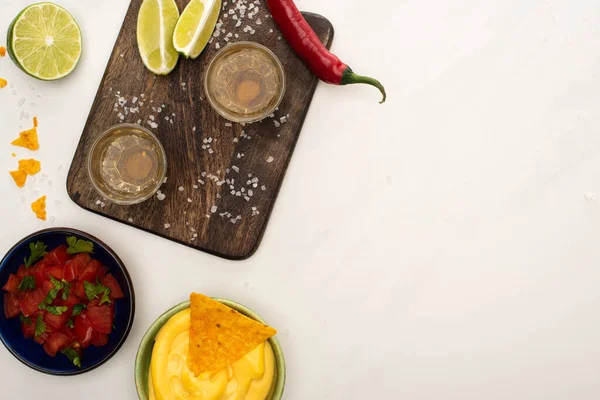 Top view of golden tequila with lime, chili pepper, salt and nachos with cheese sauce near wooden cutting board on white marble surface — Stock Photo