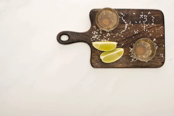 Vue de dessus de tequila dorée à la chaux, sel sur planche à découper en bois sur marbre blanc — Photo de stock