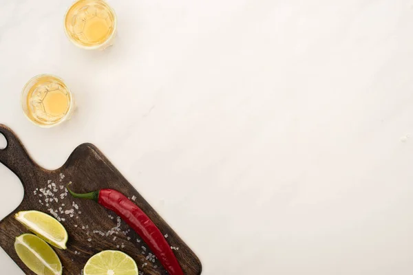 Vista dall'alto di tequila dorata con lime, peperoncino, sale su tagliere in legno su superficie di marmo bianco — Foto stock