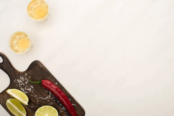 Top view of golden tequila with lime, chili pepper, salt on wooden cutting board on white marble surface — Stock Photo