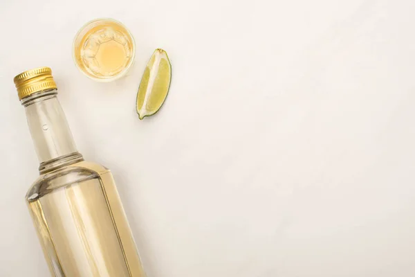 Vista dall'alto di tequila dorata in bottiglia e bicchierino con calce su superficie di marmo bianco — Foto stock