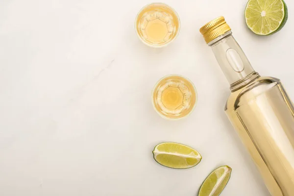 Vue de dessus de la tequila dorée en bouteille et verres de tir avec de la chaux sur la surface de marbre blanc — Photo de stock