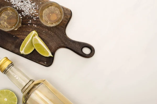 Vista superior de tequila dorado con cal, sal sobre tabla de cortar de madera sobre superficie de mármol blanco - foto de stock