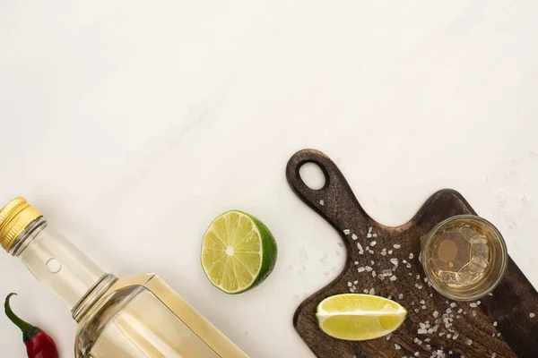 Top view of golden tequila with lime, chili pepper, salt on wooden cutting board on white marble surface — Stock Photo