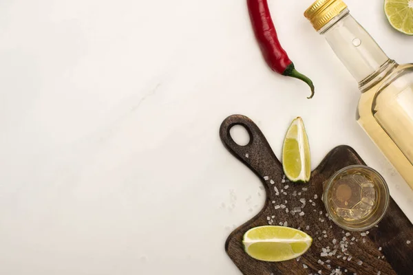 Top view of golden tequila with lime, chili pepper, salt on wooden cutting board on white marble surface — Stock Photo