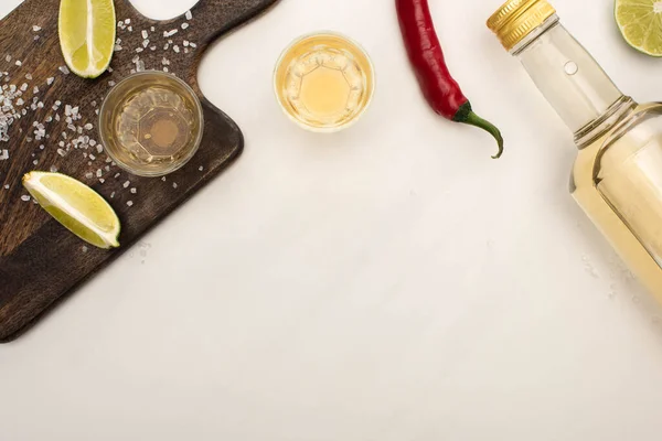 Top view of golden tequila with lime, chili pepper, salt on wooden cutting board on white marble surface — Stock Photo