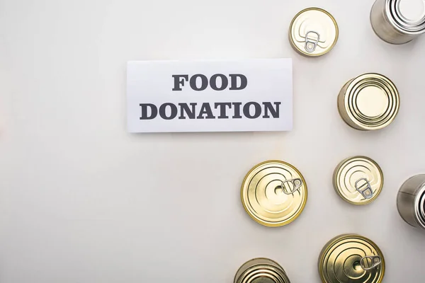 Vista superior de latas perto do cartão com letras de doação de alimentos no fundo branco — Fotografia de Stock