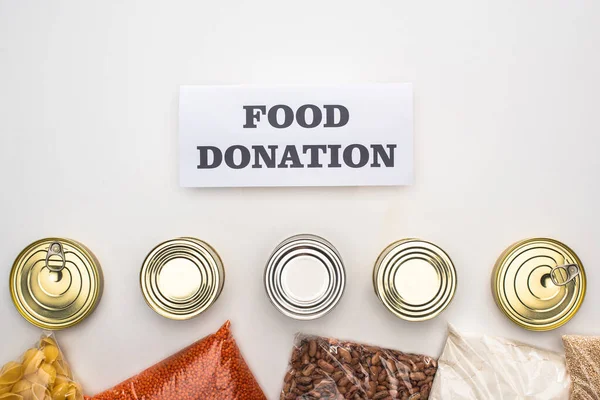 Vista superior de latas e grumos em sacos de zíper perto do cartão com letras de doação de alimentos no fundo branco — Fotografia de Stock