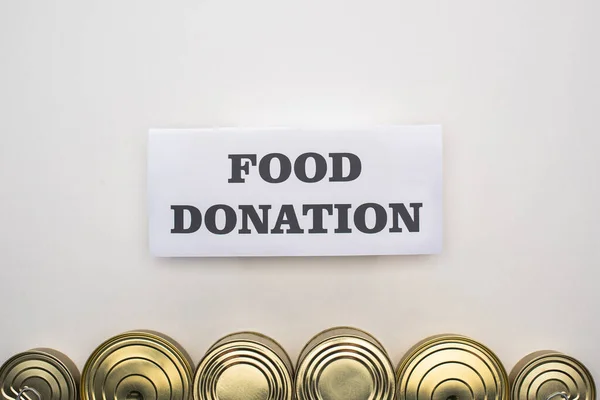 Vista superior de latas con comida enlatada sobre fondo blanco con tarjeta de donación de alimentos - foto de stock