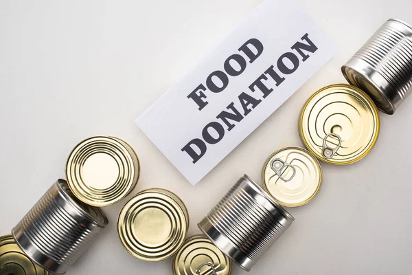 Vista superior de latas con comida enlatada sobre fondo blanco con tarjeta de donación de alimentos - foto de stock