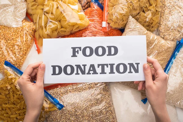 Vista recortada de la mujer que sostiene la tarjeta con letras de donación de alimentos cerca de grañones y pasta en bolsas con cremallera - foto de stock