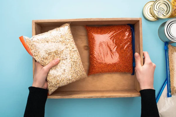 Cropped view of woman packing groats in wooden box on blue background, food donation concept — Stock Photo