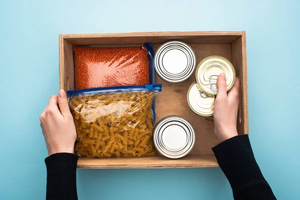 Cropped view of woman holding wooden box with cans and groats in zipper bags on blue background — Stock Photo