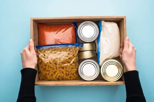 Cropped view of woman holding wooden box with cans and groats in zipper bags on blue background — Stock Photo