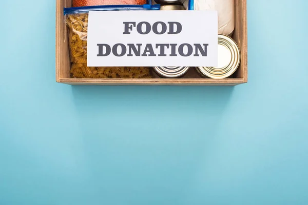 Top view of cans and groats in zipper bags in wooden box near card with food donation lettering on blue background — Stock Photo