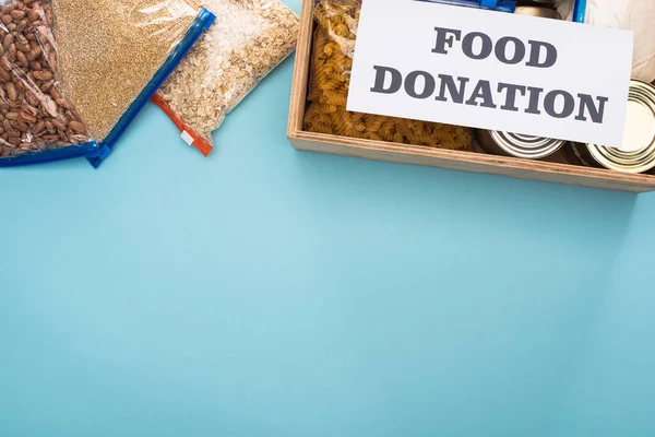 Vista superior de latas y grañones en bolsas con cremallera en caja de madera cerca de la tarjeta con letras de donación de alimentos sobre fondo azul - foto de stock