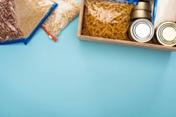 Top view of cans and groats in zipper bags in wooden box on blue background, food donation concept — Stock Photo