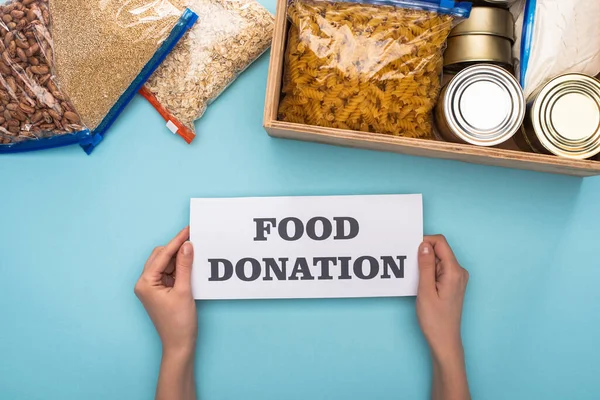 Vista recortada de la mujer que sostiene la tarjeta con letras de donación de alimentos cerca de latas y grañones en bolsas de cremallera en caja sobre fondo azul - foto de stock