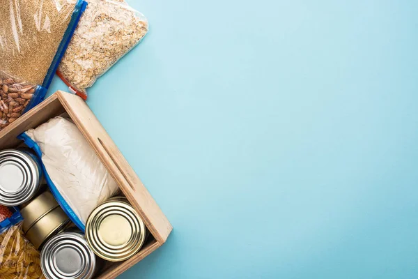 Top view of cans and groats in zipper bags in wooden box on blue background, food donation concept — Stock Photo