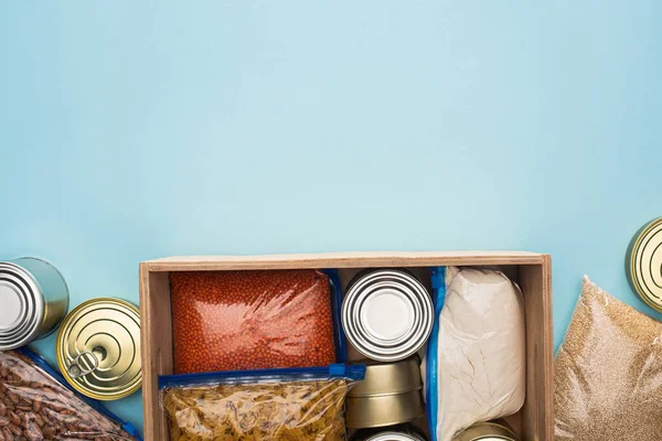 Vista superior de latas y grañones en bolsas con cremallera en caja de madera sobre fondo azul, concepto de donación de alimentos - foto de stock