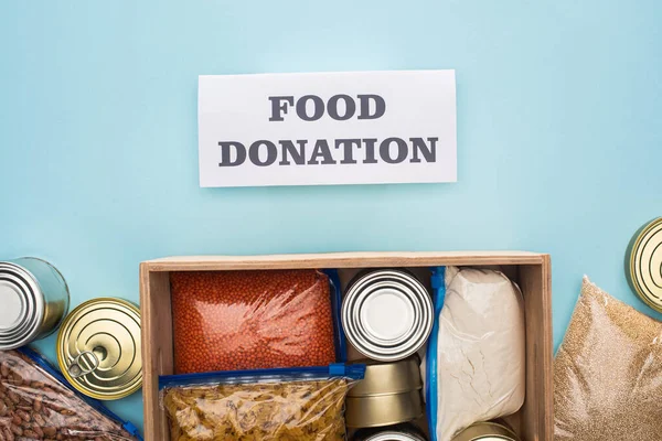 Top view of cans and groats in zipper bags in wooden box near card with food donation lettering on blue background — Stock Photo