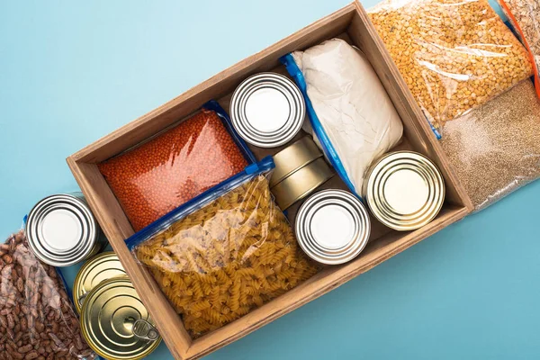 Vista superior de latas y grañones en bolsas con cremallera en caja de madera sobre fondo azul, concepto de donación de alimentos - foto de stock