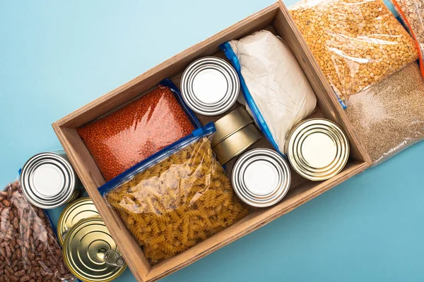 Top view of cans and groats in zipper bags in wooden box on blue background, food donation concept — Stock Photo