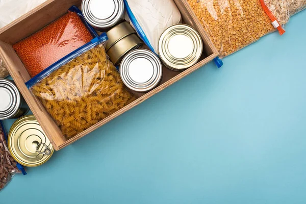 Ansicht von Dosen und Grütze in Reißverschlusstaschen in Holzkiste auf blauem Hintergrund mit Kopierraum, Konzept der Lebensmittelspende — Stockfoto