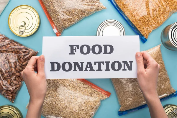 Cropped view of woman holding card with food donation lettering near cans and groats in zipper bags on blue background — Stock Photo