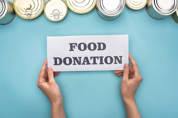 Vista cortada da mulher cartão de retenção com alimentos doação lettering perto de latas no fundo azul — Fotografia de Stock