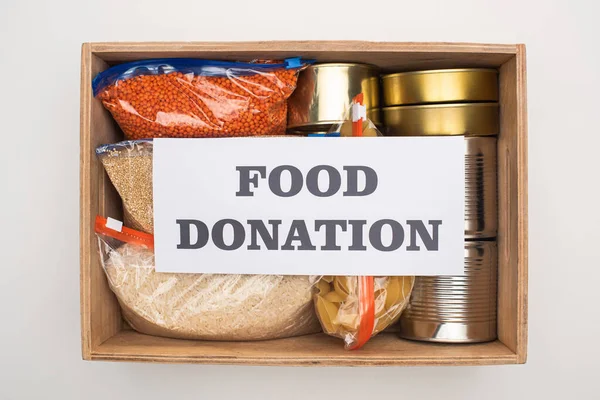 Vista superior de latas y grañones en bolsas con cremallera en caja de madera con tarjeta de donación de alimentos sobre fondo blanco - foto de stock