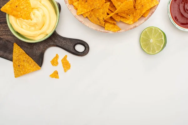 Top view of corn nachos with lime, ketchup, cheese sauce on wooden cutting board on white background — Stock Photo