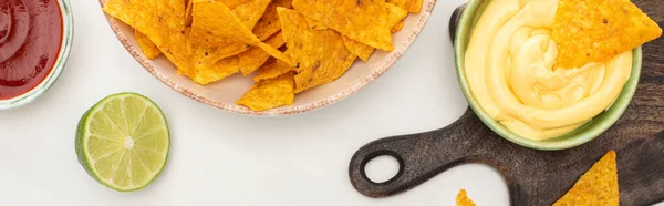 Top view of corn nachos with lime, ketchup, cheese sauce on wooden cutting board on white background, panoramic shot — Stock Photo