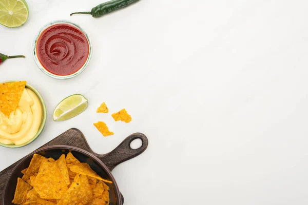 Vista dall'alto di nachos di mais con lime, peperoncino, ketchup e salsa di formaggio su tagliere di legno su sfondo bianco — Foto stock