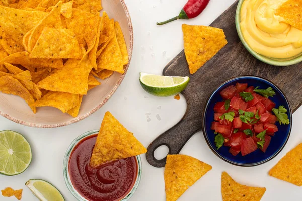 Vista superior de nachos de maíz con salsa de lima, chile, ketchup y queso sobre tabla de cortar de madera sobre fondo blanco - foto de stock