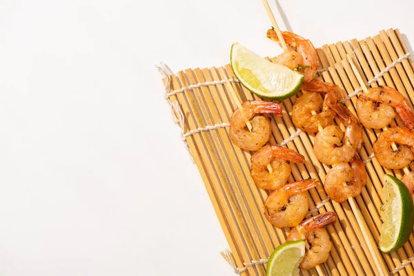 Vue de dessus des crevettes sur les brochettes avec de la chaux sur le tapis de bambou sur fond blanc — Photo de stock