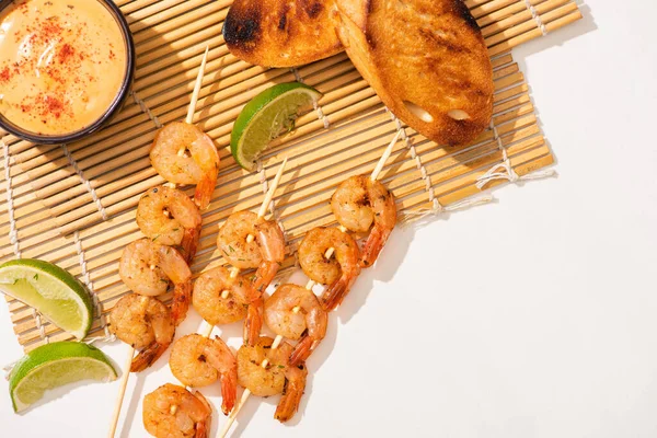 Vista superior de langostinos sobre brochetas con tostadas, lima y salsa sobre estera de bambú sobre fondo blanco - foto de stock