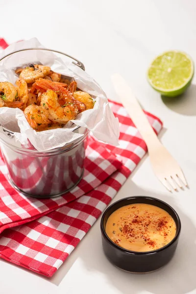 Selective focus of prawns on skewers on metal bucket and plaid napkin near lime, fork,  and sauce on white background — Stock Photo