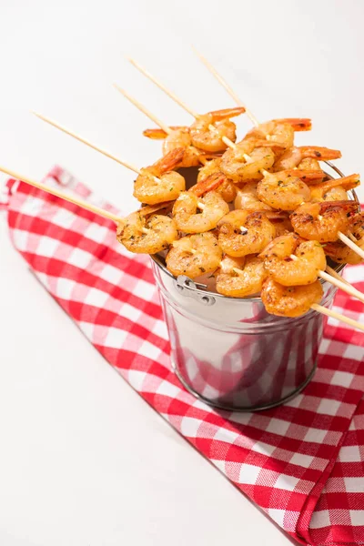 Crevettes sur brochettes sur seau en métal et serviette à carreaux sur fond blanc — Photo de stock