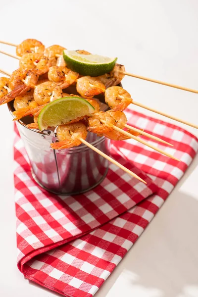 Prawns on skewers with lime on metal bucket and plaid napkin on white background — Stock Photo