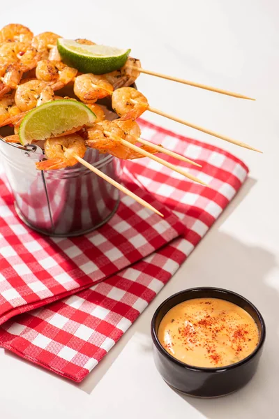 Prawns on skewers with lime on metal bucket and plaid napkin near sauce on white background — Stock Photo
