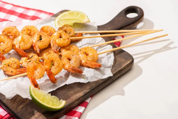 Prawns on skewers with lime on parchment paper on wooden board and plaid napkin on white background — Stock Photo