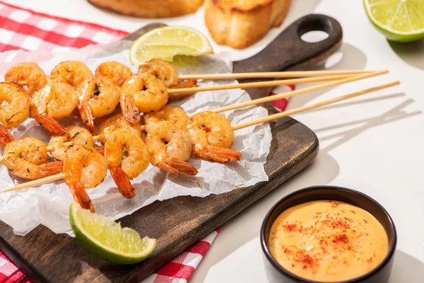 Prawns on skewers with lime and sauce on parchment paper on wooden board and plaid napkin on white background — Stock Photo