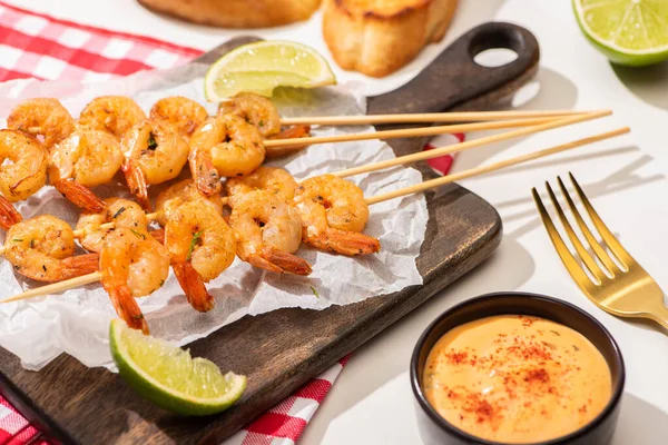 Prawns on skewers with lime and sauce on parchment paper on wooden board and plaid napkin on white background — Stock Photo