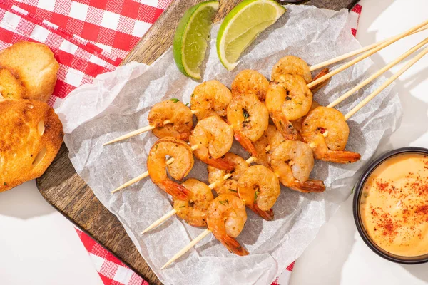 Prawns on skewers with lime, sauce, bread toasts on parchment paper on wooden board and plaid napkin on white background — Stock Photo