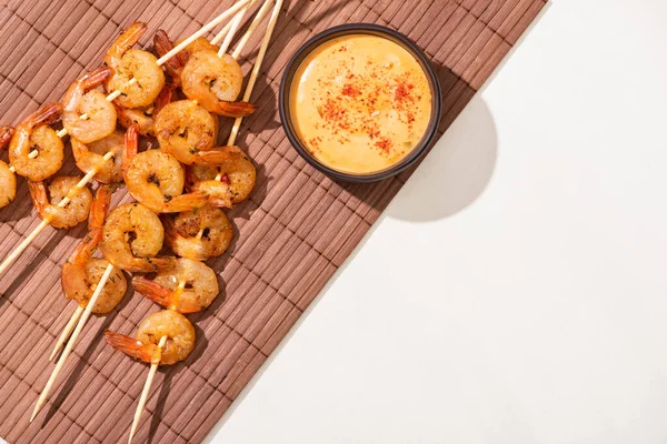 Vista superior de sabrosos langostinos en brochetas con salsa sobre estera de bambú sobre fondo blanco - foto de stock